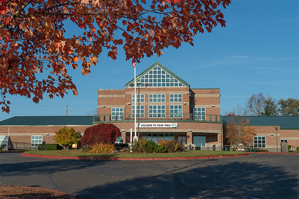 A brick school building with a green roof, surrounded by trees with red leaves. A flagpole stands in front. A banner reads,