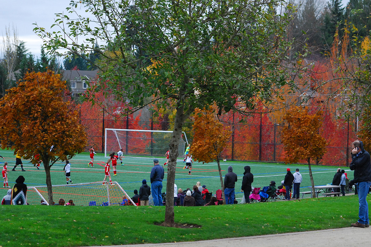 People watch a youth soccer game on a field surrounded by autumn trees, with a man taking photos on the sideline.