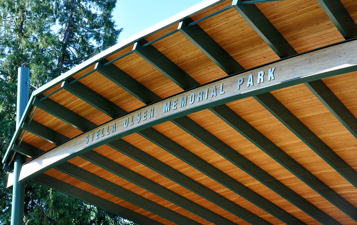 A wooden park shelter with a curved roof displaying the name "Stella Olsen Memorial Park" in large letters.