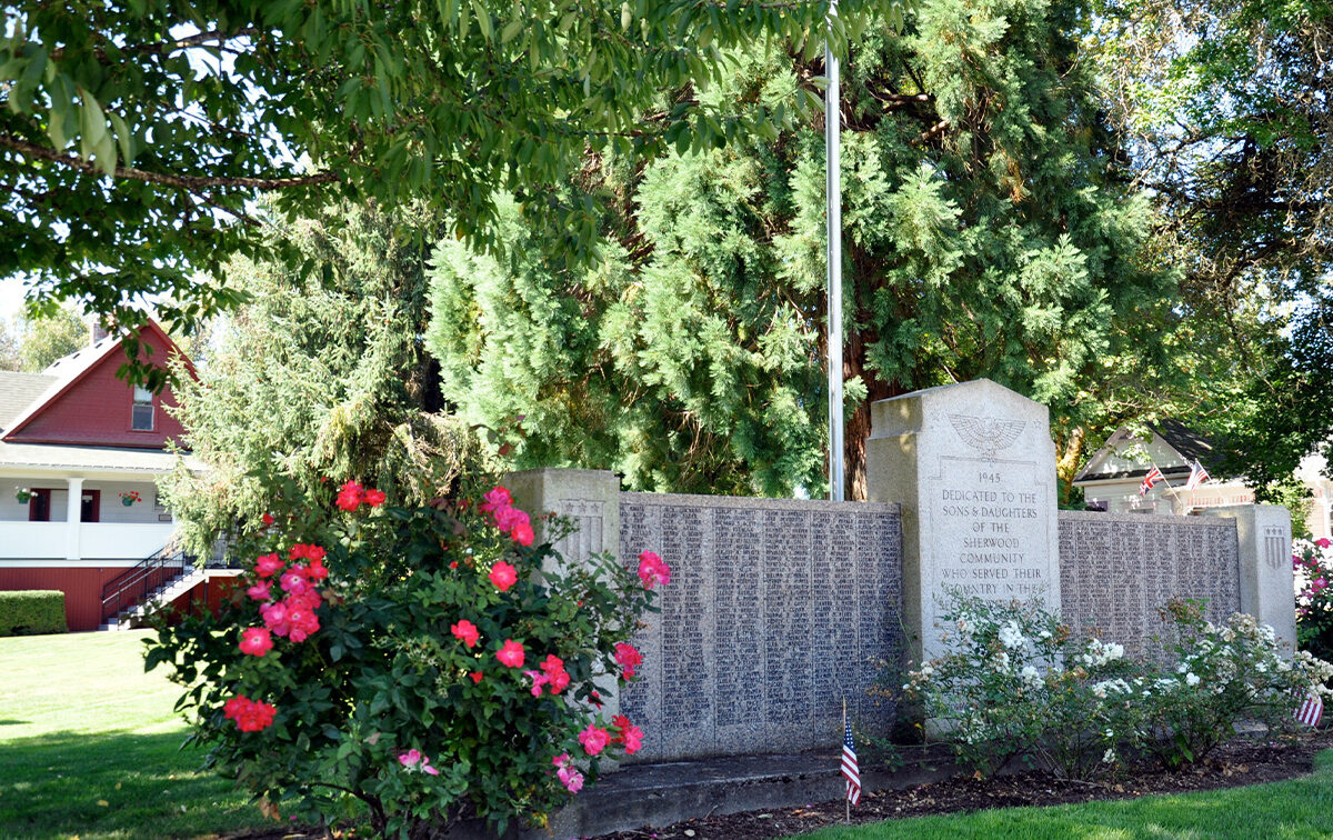 A memorial wall with engraved names stands under trees, surrounded by flowering bushes and small flags. A red-roofed house is visible in the background.