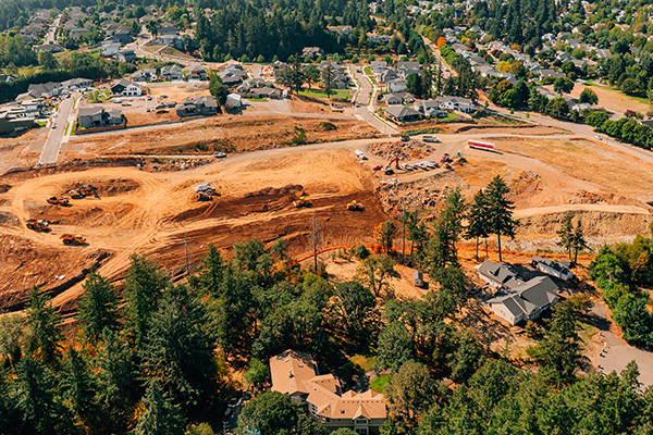 Aerial view of a residential neighborhood under construction, with cleared land, construction vehicles, and nearby houses surrounded by trees.