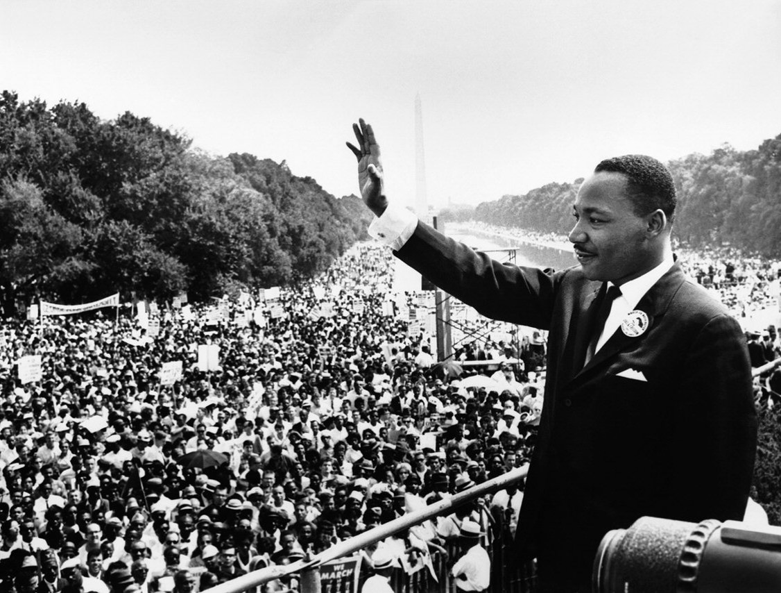 A man in a suit stands on a platform, waving to a large crowd gathered outdoors with trees lining the background.